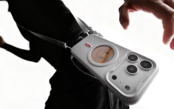A person wearing black carries a white crossbody bag shaped like a smartphone with camera lenses and a small round screen displaying a cartoon face.